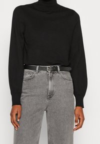 Schwarzer Rollkragenpullover mit langen Ärmeln, kombiniert mit hoch taillierten grauen Jeans und einem schwarzen Ledergürtel. Subtile Textur und taillierte Passform.