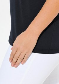 Top de tela suave y negra con un dobladillo redondeado, combinado con jeans ajustados blancos. Una mano descansa suavemente en el lado del top, mostrando la textura.