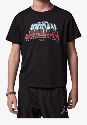 Barn iført en sort Air Jordan t-shirt med rødt, hvidt og blåt skrift samt sorte bukser med et hvidt Jumpman-logo på venstre ben.