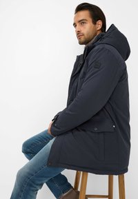 Man in een donkere winterjas met capuchon en blauwe jeans, zijwaarts zittend op een houten kruk tegen een witte achtergrond.