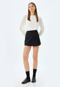 Blusa bianca a maniche lunghe con maniche trasparenti plissettate, abbinata a una mini gonna nera plissettata. Mocassini neri e calze completano il look.