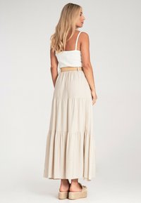 Beige, schichtiger Maxirock mit strukturiertem Finish, kombiniert mit einem enganliegenden weißen Tanktop. Das Modell steht seitlich und zeigt den Bund sowie die Schuhe.