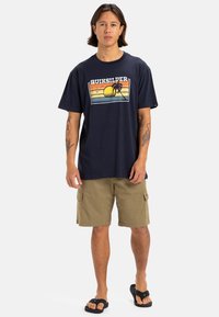 Camiseta navy con estampado gráfico que presenta un atardecer y palmeras; combinada con pantalones cortos khaki y sandalias negras. Corte casual, mangas cortas.