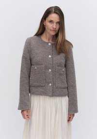 Ruskea teksturoitu villan sekoitekangas takki, jossa on pyöreä kaulus, edessä nappisuljenta ja kaksi päystä taskua. Pitkät hihat viimeistelevät muotoilun.