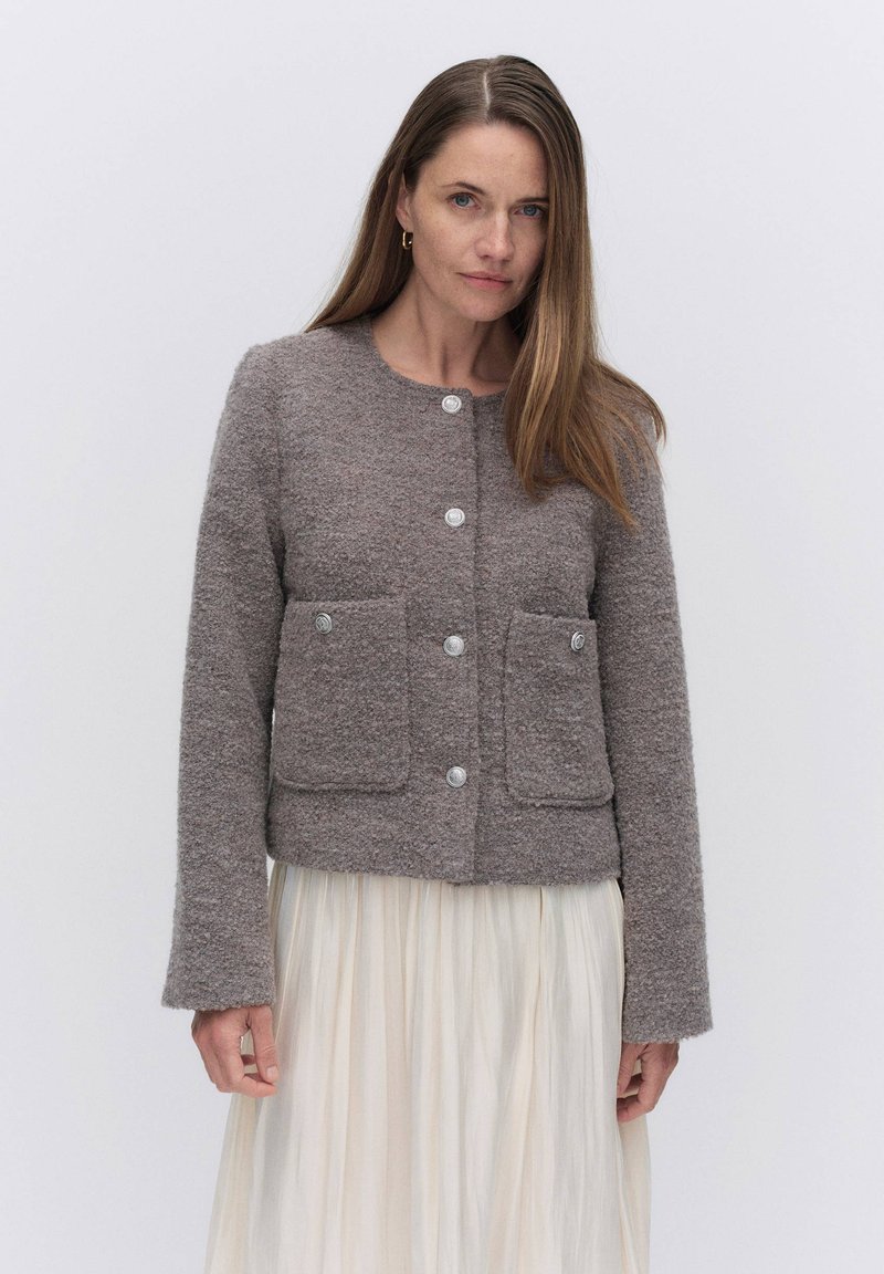 Ruskea teksturoitu villan sekoitekangas takki, jossa on pyöreä kaulus, edessä nappisuljenta ja kaksi päystä taskua. Pitkät hihat viimeistelevät muotoilun.