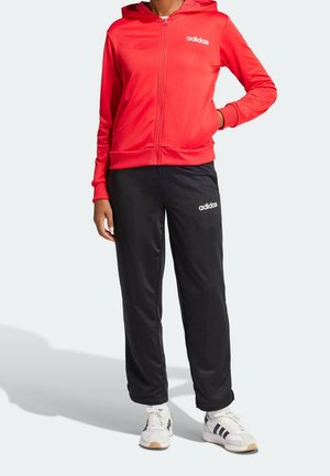 Person trägt eine rote Adidas Zip-up-Hoodie, schwarze Adidas Trainingshose und weiße Adidas Sneakers mit schwarzen Streifen, steht vor einem hellen Hintergrund.