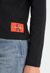 Chemise noire à manches longues avec une étiquette logo Calvin Klein Jeans en orange, associée à un jean en denim bleu. Gros plan sur l'ourlet et la manche de la chemise.