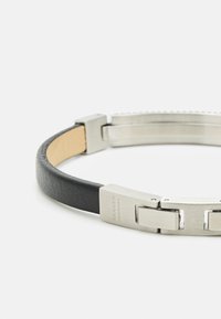 Bracciale in pelle nera con chiusura in acciaio inox argentato. Presenta una texture liscia e un design a catena, incisa con "SKAGEN DENMARK".