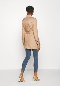 Beżowy trencz z paskiem, z wyraźnie zarysowanymi ramionami i zapinanymi mankietami, zestawiony z przetartymi niebieskimi rurkami i białymi sandałami na obcasie.