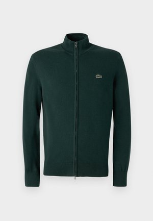 Sweat-shirt zippé vert foncé avec un col haut, des poignets et un ourlet côtelés. Il présente un petit logo brodé sur la poitrine. Texture de tissu douce.