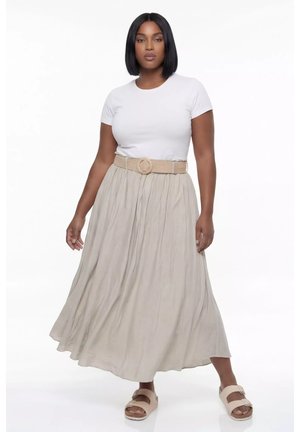 Kobieta o prostych czarnych włosach, ubrana w biały top z krótkim rękawem, beżową plisowaną długą spódnicę z paskiem oraz beżowe sandały, stojąca na białym tle.