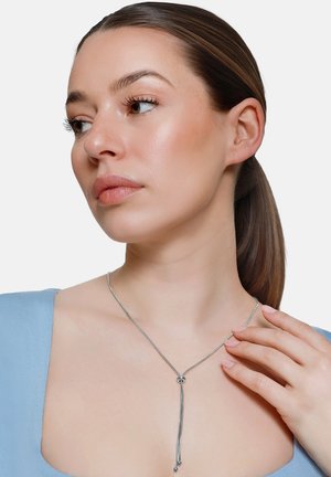 Kobieta z brązowymi włosami związanymi w kucyk, ubrana w jasnoniebieski top i srebrny naszyjnik lariat, dotykająca naszyjnika prawą ręką.