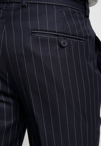 Marinblå ränderade byxor med en enda bakficka, featuring en knappstängning och en slät tygtextur.