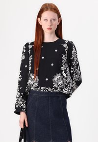 Schwarze Bluse mit weißen Blumenmustern, langen Ärmeln, runden Ausschnitt, kombiniert mit einem Jeansrock mit Zickzacksaum.