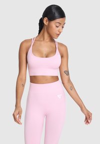 Ženska z zapletenimi lasmi, ki nosi roza športni modrček in hlače z visokim pasom, gleda navzdol z sproščenimi rokami, tetovirana na obeh rokah.