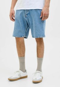 Bermudas de mezclilla en azul claro, de corte ligeramente suelto, con dobladillo deshilachado, usadas con calcetines grises y zapatillas blancas con detalles en gris y suelas de goma.