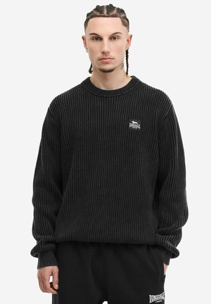 Ung mand med flettet hår iført sort ribbet sweater og sorte bukser, begge med Lonsdale London-logo, foran en ensfarvet baggrund.