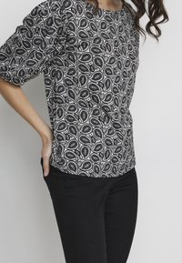 Una persona indossa una maglia a maniche puff con motivo paisley in bianco e nero, abbinata a pantaloni neri, con le mani infilate nelle tasche.