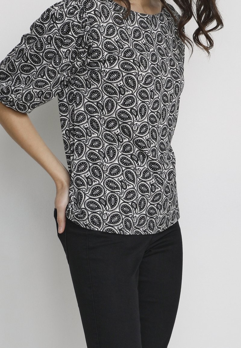 Una persona indossa una maglia a maniche puff con motivo paisley in bianco e nero, abbinata a pantaloni neri, con le mani infilate nelle tasche.