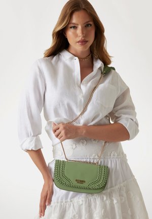 Borsa a tracolla verde in ecopelle con texture intrecciata, tracolla in catena dorata e dettaglio del logo. Indossata con una camicia bianca a maniche lunghe e una gonna.