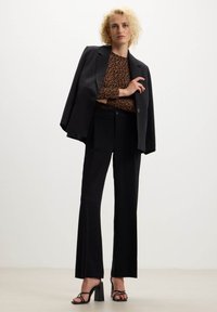 Blazer nero sopra un top marrone con stampa leopardata, abbinato a pantaloni neri a vita alta e tacchi a strappo neri. Tessuto liscio e vestibilità su misura.