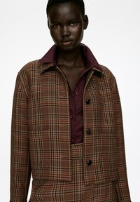 Brown plaid cropped jacket with black buttons, featuring a maroon collar. Worn over a maroon shirt with contrasting buttons. Textured fabric.