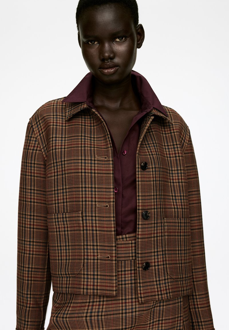Brown plaid cropped jacket with black buttons, featuring a maroon collar. Worn over a maroon shirt with contrasting buttons. Textured fabric.