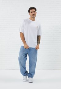 Weißes T-Shirt mit kurzen Ärmeln und einem kleinen bunten Grafikdesign auf der linken Brust, kombiniert mit lockeren blauen Jeans und weißen Turnschuhen.