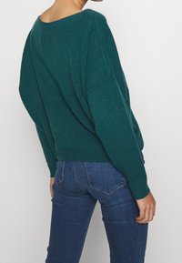 Türkiser Strickpullover mit weitem Ausschnitt und überschnittenen Schultern. Kombiniert mit dunkelblauen, enganliegenden Jeans, die eine glatte Textur und eine lässige Passform aufweisen.