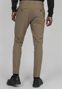 Pantalons ajustés marron clair avec une finition texturée, dotés de deux poches arrière, associés à des chaussures noires. Retroussés aux chevilles.