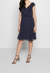 Navy blue polka dot dress with ruffled neckline, short sleeves, and flared hem. Lightweight fabric, knee-length cut, paired with black strappy heels.