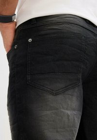 Jean en denim noir avec une texture légèrement délavée, présentant un design de poche arrière et des accents de bouton en métal sur la taille.