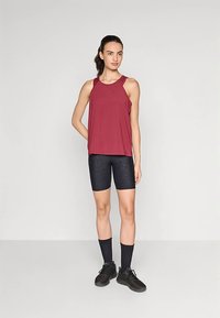 Dunkelrotes ärmelloses Oberteil kombiniert mit schwarzen, strukturierten Radlerhosen und schwarzen Turnschuhen. Das Model steht vor einem schlichten Hintergrund.