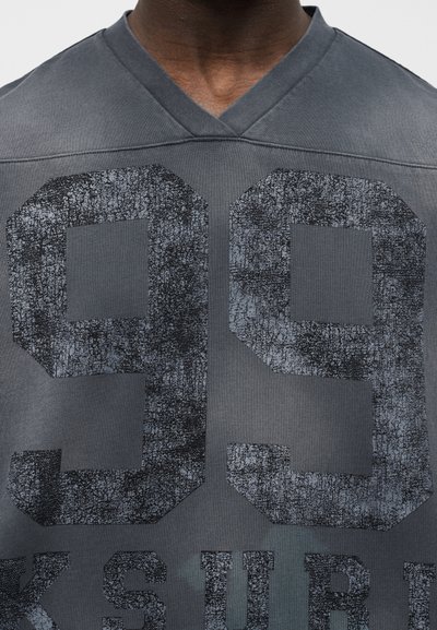 Κοντινό πλάνο ενός ατόμου που φοράει γκρι μπλουζάκι με V λαιμόκοψη και μεγάλο, φθαρμένο μαύρο αριθμό 99 τυπωμένο μπροστά.