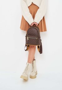 Zaino in pelle marrone con motivo esagonale, cerniere dorate, forma arrotondata e spallacci regolabili, abbinato a stivali beige con lacci.