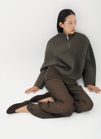 Γυναίκα με μαύρα μαλλιά που φοράει σκούρο γκρι μπουφάν με φερμουάρ, διαφανές καφέ παντελόνι και καφέ φλατ παπούτσια, καθισμένη σε λευκό δάπεδο δίπλα σε τοίχο.