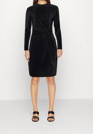 Kobieta ubrana w czarną welurową sukienkę z długim rękawem, sięgającą do kolan, z marszczeniem w talii, oraz czarne sandały na obcasie z odkrytymi palcami i paskiem.