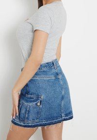 Femme portant un haut ajusté à manches courtes gris clair et une mini-jupe en jean bleue avec une poche latérale et un ourlet effiloché.