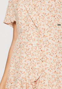 Robe légère à motifs floraux dans des tons pêche doux avec un ourlet à volants, ornée de grands boutons clairs et de manches courtes. Le matériau semble texturé.