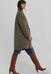 Brun og beige houndstooth-frakke, mellemlang, med krave og sidelommer, parret med tætsiddende brune knæhøje støvler og blå bukser.