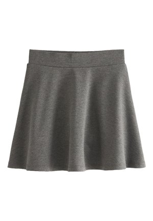 Enfant portant une chemise blanche, une jupe grise, des chaussettes blanches et des chaussures noires debout à l'intérieur avec des papiers roses et des crayons au sol à proximité.