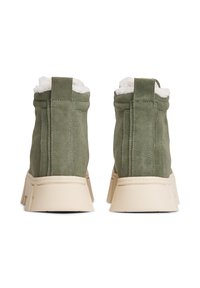 Une paire de bottes d'hiver en daim vert avec une doublure en fausse fourrure blanche, des semelles épaisses beiges, vues de l'arrière sur un fond blanc.