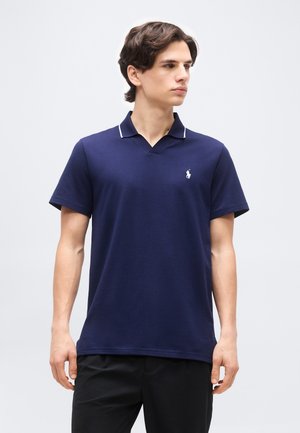 Νεαρός άνδρας με μπλε ναυτικό κοντομάνικο πόλο με λευκό λογότυπο, όρθιος και κοιτάζει προς τα δεξιά, με φόντο απλό ανοιχτό γκρι.