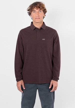 Homme aux cheveux bouclés portant un polo marron foncé à manches longues et un pantalon gris, debout devant un fond clair uni.