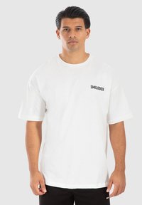 Hvid bomulds t-shirt med korte ærmer, rund hals og "SMILODOX" logo i sort på venstre brystområde. Enkel design, afslappet pasform.