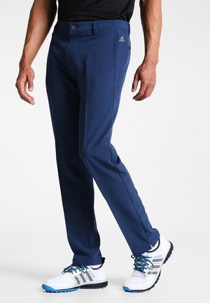 Uomo con pantaloni Adidas blu navy e scarpe da ginnastica bianche con lacci blu, in piedi con le mani rilassate ai lati.