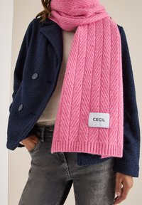 Rosa gerippter Strickschal mit Fischgrätenmuster und einem weißen Etikett, auf dem "CECIL" steht, getragen mit einer marineblauen Fleecejacke und grauen Hosen.