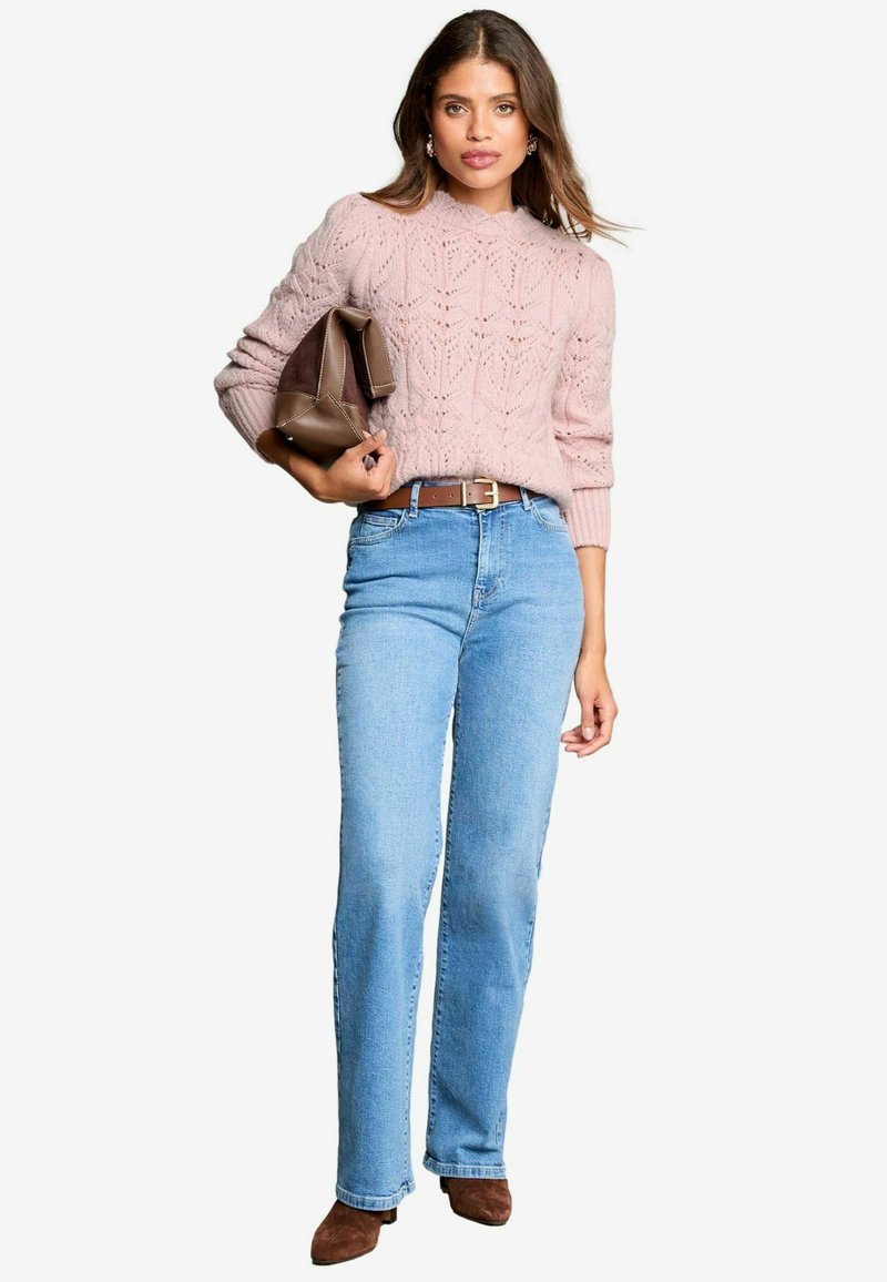 Maglione lavorato a maglia rosa chiaro con un motivo texturizzato, abbinato a jeans dritti blu medio e stivali ankle marroni; tiene una borsa marrone.