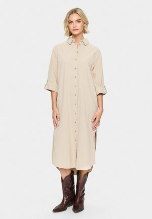 Beige knoopshirt jurk met opgerolde mouwen, twee zijslitsen en subtiele textuur, gecombineerd met donkerbruine cowboylaarzen.