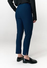 Marineblauwe stretchbroek met een getailleerde pasvorm, gespleten zoom bij de enkel en een gladde textuur, gecombineerd met glanzende zwarte loafers.